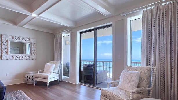 Ein geräumiges Wohnzimmer mit einer großen Glasschiebetür, die auf einen Balkon mit Meerblick führt. Zwei weiße, gepolsterte Sessel und ein kleiner weißer Hocker sind ebenfalls vorhanden.