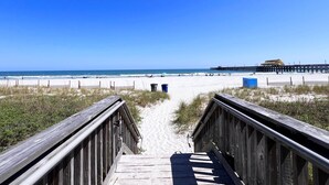 Ein hölzerner Steg führt zu einem Strand, im Hintergrund ist ein Steg zu sehen.