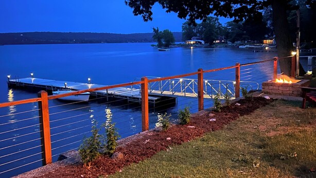Un muelle se adentra en un lago tranquilo al anochecer, con una zona cubierta de césped y un árbol cercano.