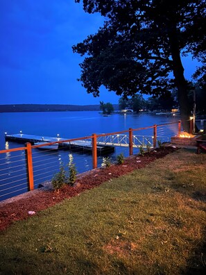 Un muelle se adentra en un lago tranquilo al anochecer, con una zona cubierta de césped y un árbol cercano.
