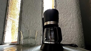 Une machine à café posée sur un sol carrelé, près d’une fenêtre, avec un mur à la texture particulière.
