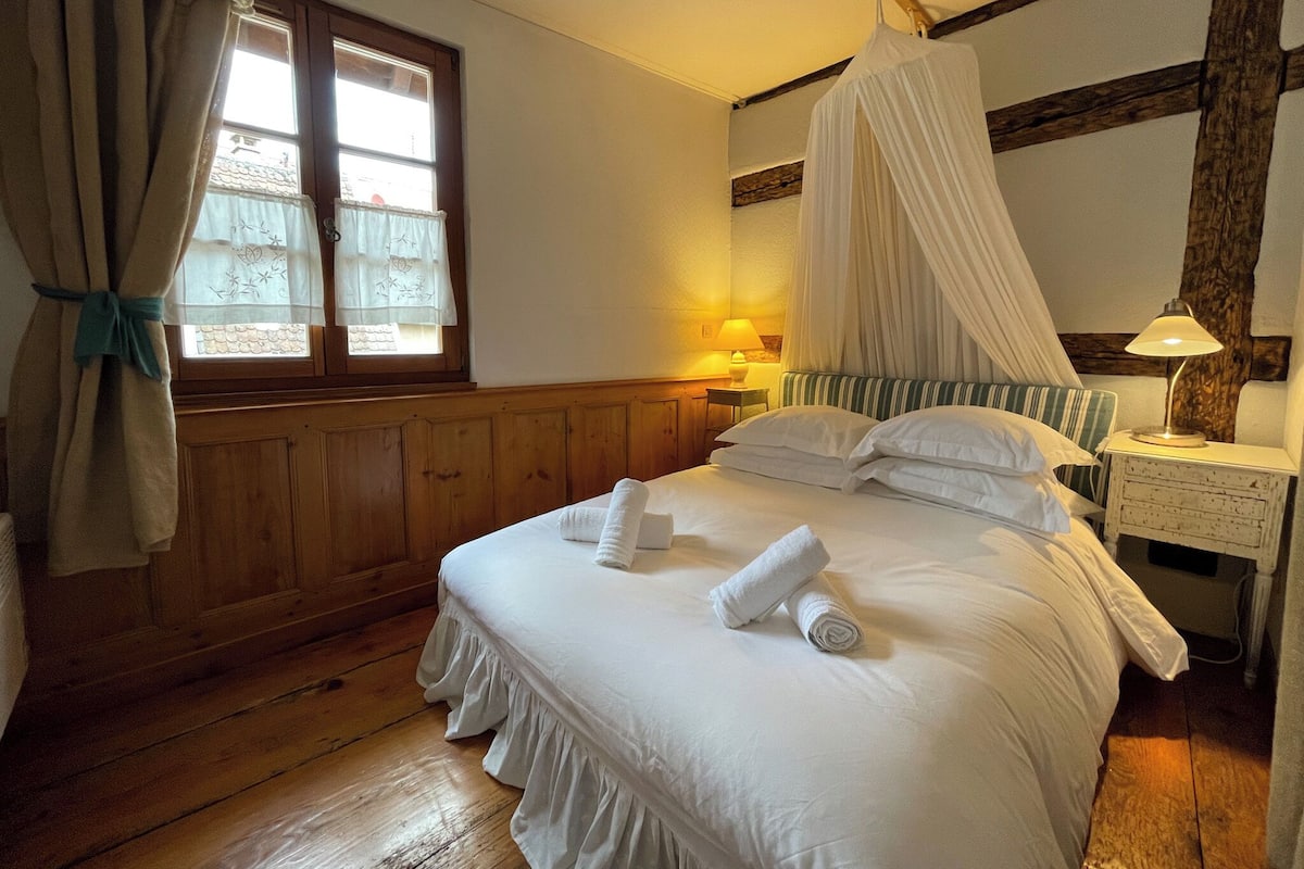 A bedroom with a four-poster bed, a bedside table with a lamp, and a window with curtains.