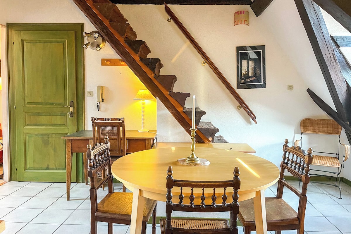 A dining area with a round table, wooden chairs, a green door, a staircase, and a wall-mounted picture.