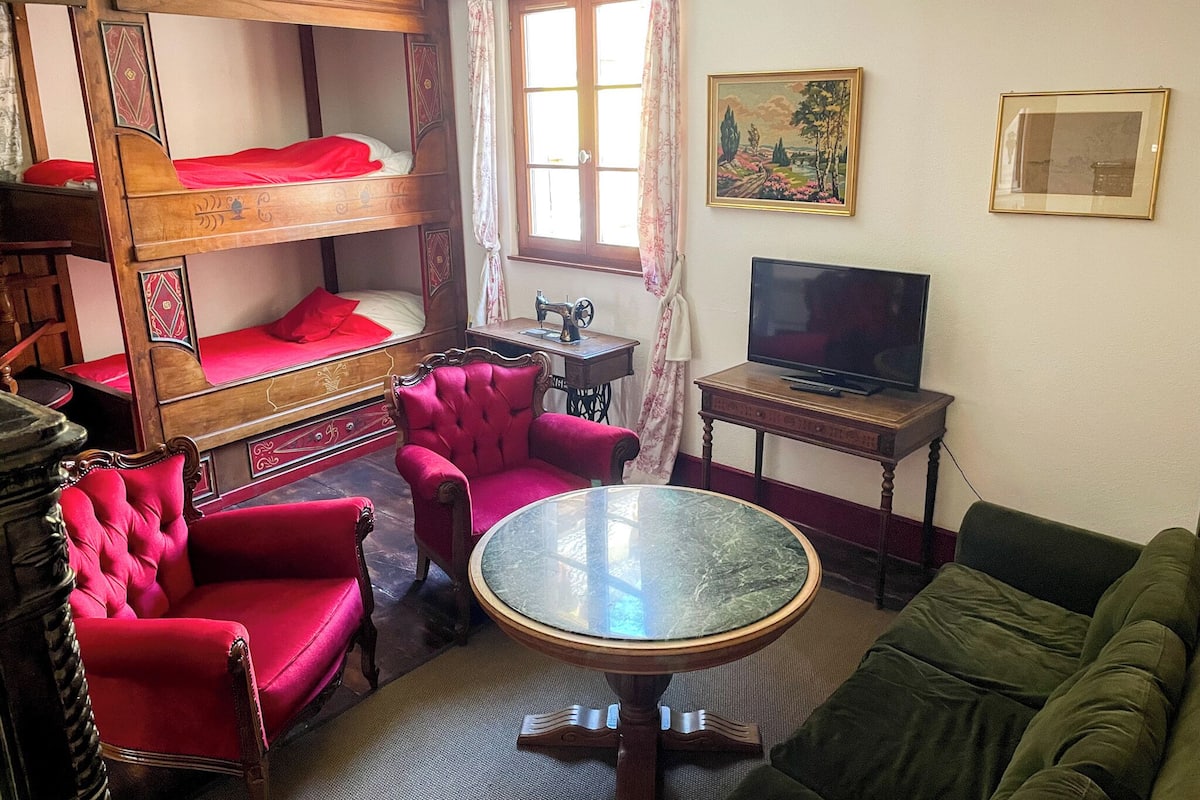 A room with a bunk bed, a red armchair, a green sofa, a round table, a television, and a window with curtains.