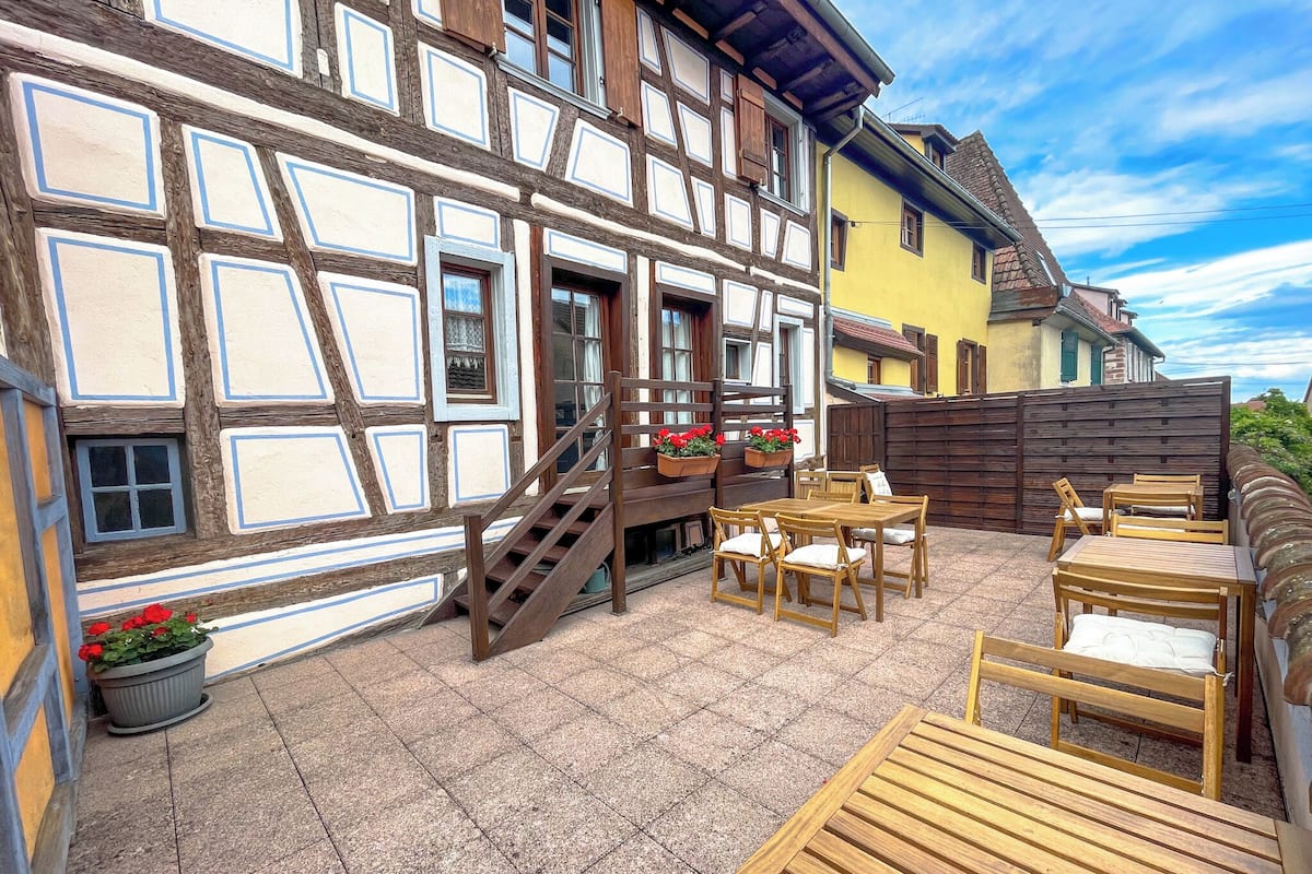 A traditional half-timbered building with a patio area featuring wooden tables and chairs, a staircase, and flower boxes.
