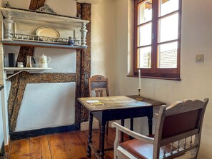 A cozy room with a wooden floor, a table with a chair, a shelf with dishes, a window with curtains, and a wall-mounted light switch. - La Maison Bleue (Riquewihr)