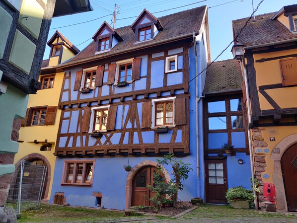 A traditional half-timbered house with a blue facade, multiple windows with wooden shutters, and a small garden area with a tree and potted plants. - La Maison Bleue (Riquewihr)