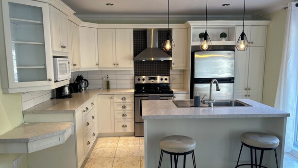 A modern kitchen with white cabinetry, a central island with stools, stainless steel appliances, and pendant lighting. - Magnifique maison près de l'eau (Laval)