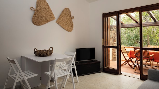 A room with a white table, two white chairs, a woven basket, a flat-screen TV, and a sliding glass door leading to an outdoor seating area with orange chairs. - Casa Girgolu by Wonderful Italy (San Teodoro)