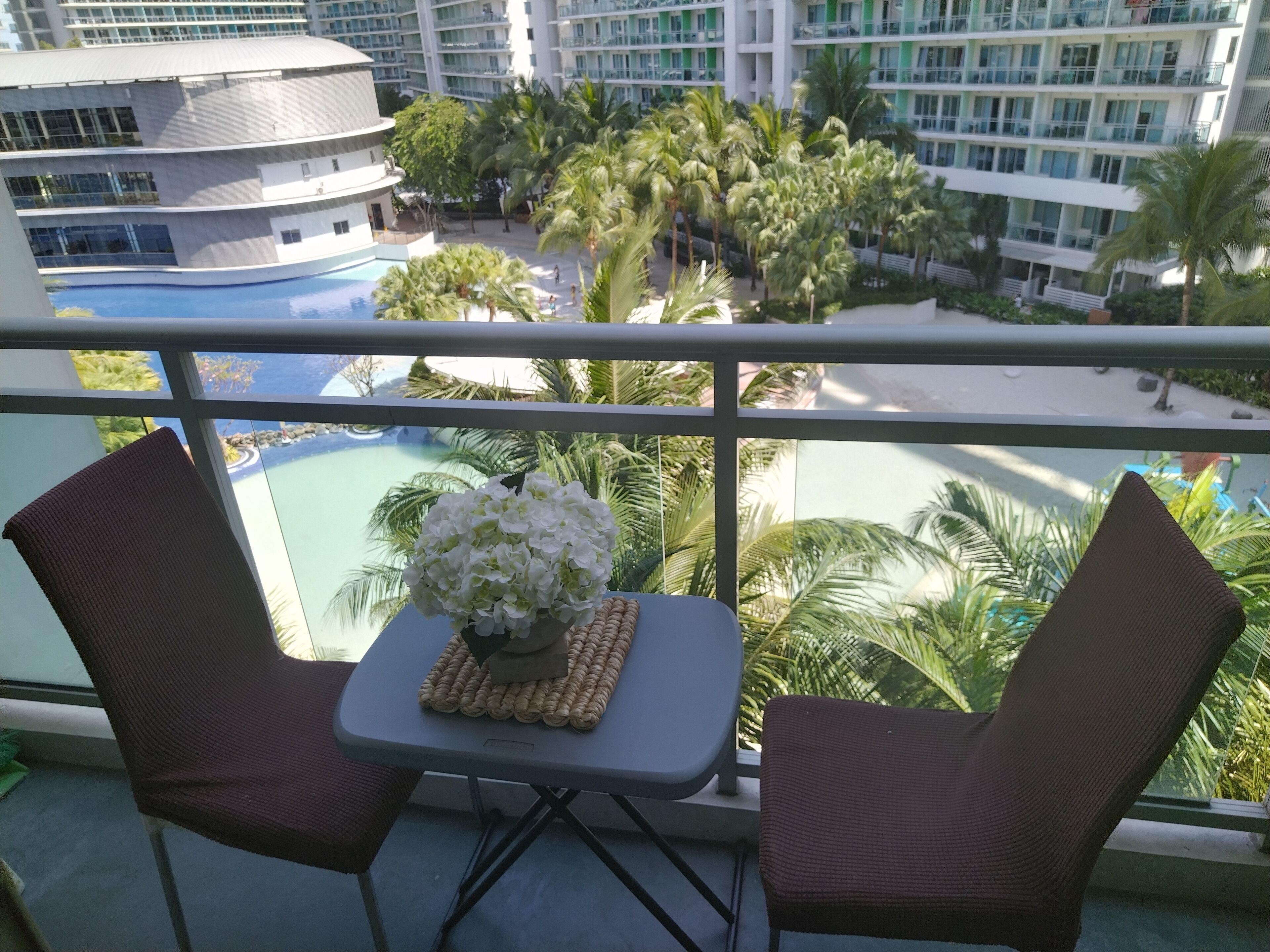 A balcony with two wicker chairs, a small table with a vase of white flowers, and a view of a pool and palm trees.