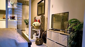 A modern living room with a sofa, a TV, a dining table, and a kitchen area visible through a glass door. - Santorini Suites (Parañaque)