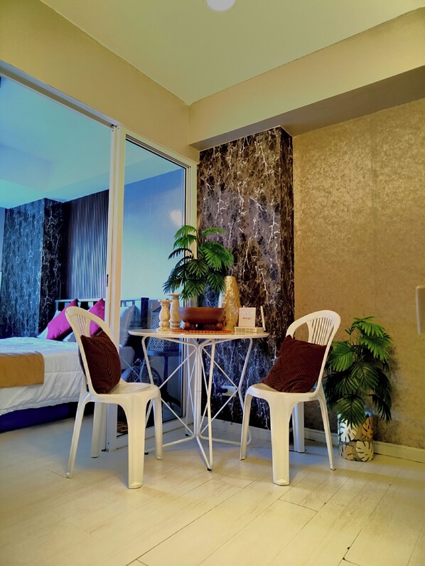 A modern room with a bed, two white chairs, a small table with a plant, and a large window. - Santorini Suites (Parañaque)