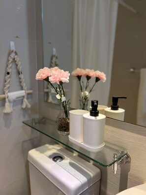 Une salle de bain avec une étagère en verre sur laquelle sont posés un vase rempli de fleurs roses, une bouteille blanche et un distributeur noir à pompe.