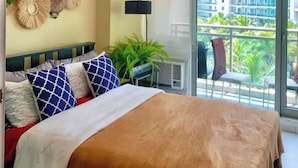 A bedroom with a bed, a wall-mounted decorative item, a floor lamp, and a view of a balcony with outdoor furniture. - Santorini Suites (Parañaque)