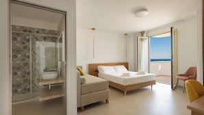 Habitación de hotel moderna con cama, sofá, una mesita, una silla, baño con ducha de vidrio y balcón con vistas.