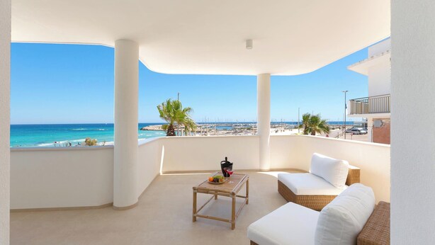 Un balcón con vistas al mar, un sofá de mimbre, una mesita con una bebida y una pared blanca.