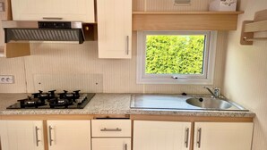 Une cuisine compacte équipée d’une cuisinière à gaz, d’une fenêtre donnant sur un espace verdoyant, d’un évier et de meubles de rangement blancs.