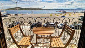 A balcony with a wooden table and chairs overlooking a beach with parked cars and boats.