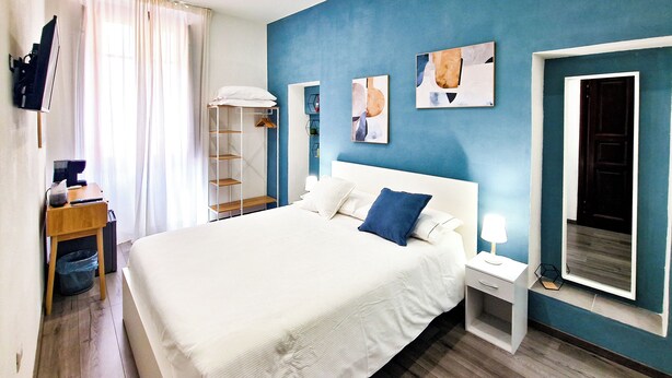 A modern hotel room with a large bed, a bedside table with a lamp, a wardrobe, and a desk with a computer.