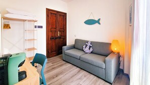 A living room with a grey sofa, wooden floor, a wooden door, a fish wall decoration, a lamp, and a small table with a printer.