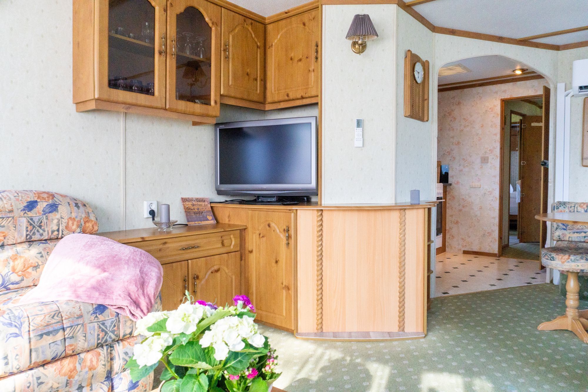 A compact living space with wooden cabinetry, a flat-screen TV, a floral sofa, and a small table with a lamp.