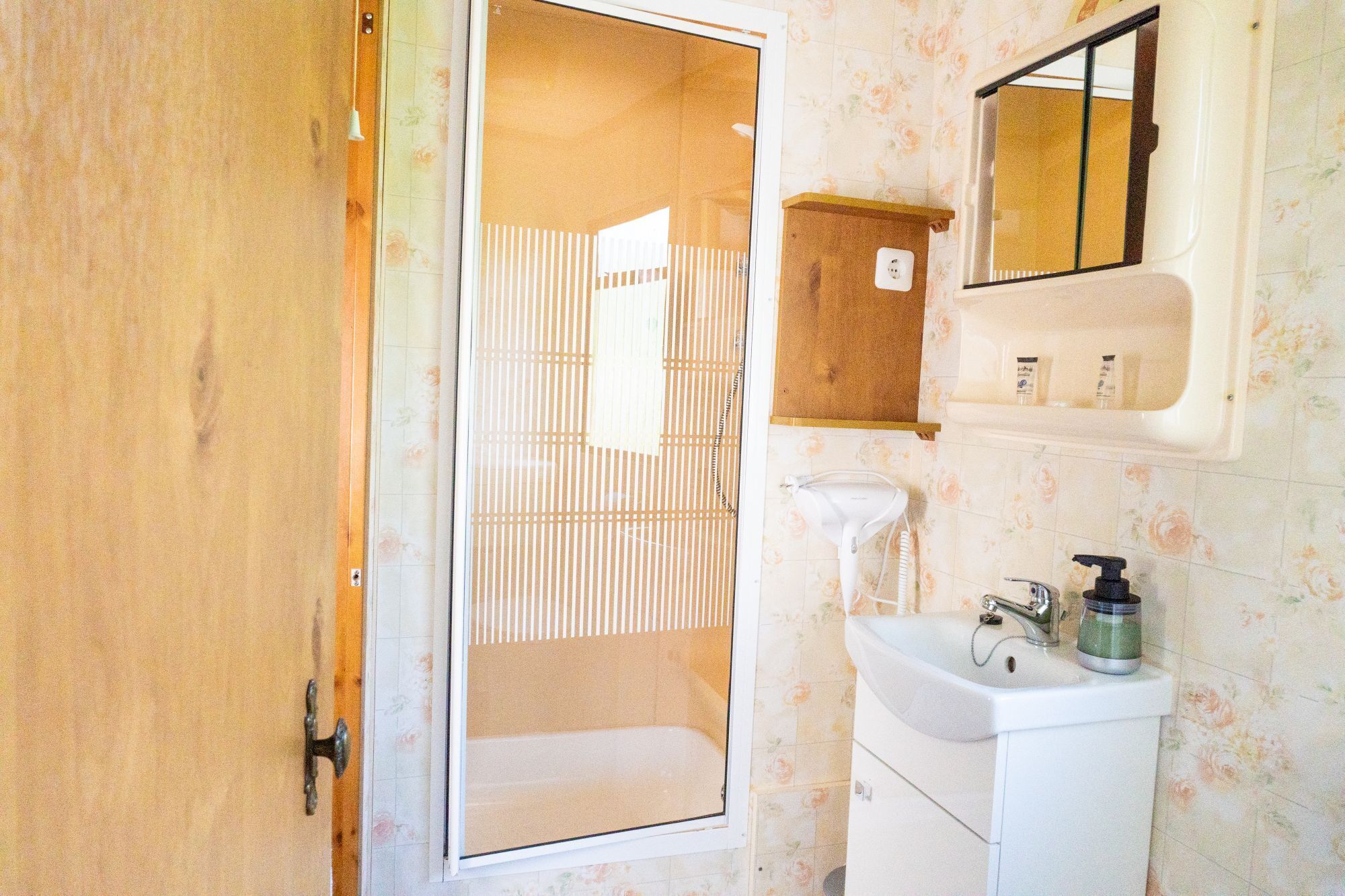 A bathroom with a wooden door, a shower, a sink with a mirror, and toiletries on the wall.