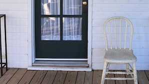 Una terraza de madera con una silla blanca y una puerta verde oscuro con cuatro ventanas.