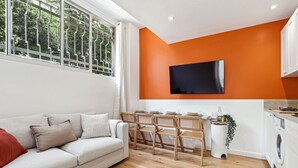 Ein modernes Wohnzimmer mit einer Couch, einem Esstisch, Stühlen, einem an einer orangefarbenen Wand montierten Fernseher und einer Waschmaschine in der Ecke.