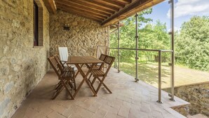 Un patio con paredes de piedra, una mesa y sillas de madera, ventanas amplias y vistas a un paisaje verde.