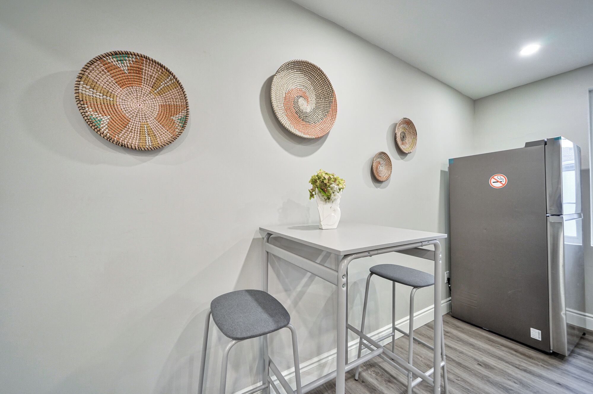 A modern kitchen with a white table, two bar stools, a refrigerator, and three woven wall hangings.