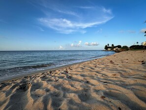 Ein sandiger Strand mit Fußspuren, die zum Wasser führen, ein klarer Himmel und ein entferntes Gebäude mit Palmen.
