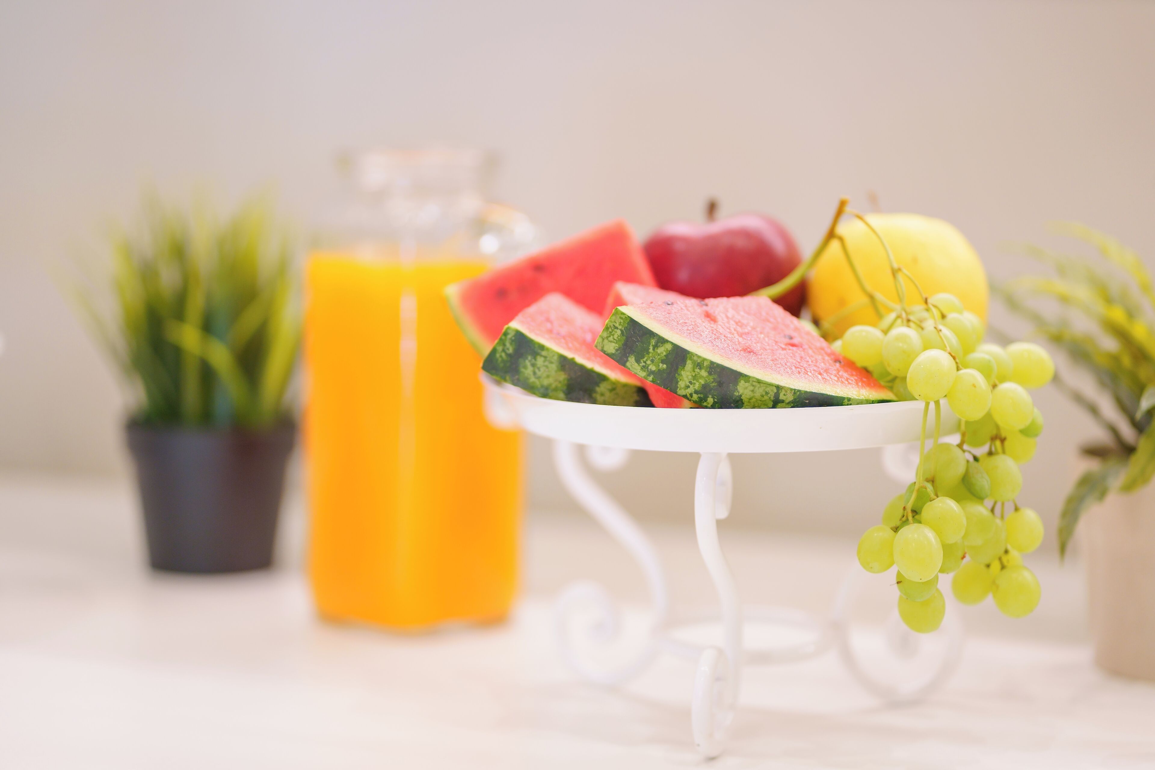 A table with a white stand holding sliced watermelon, a bunch of grapes, and a yellow fruit, with a glass of orange juice and a potted plant in the background.