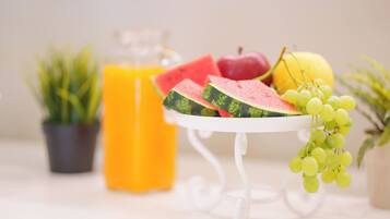 A table with a white stand holding sliced watermelon, a bunch of grapes, and a yellow fruit, with a glass of orange juice and a potted plant in the background.
