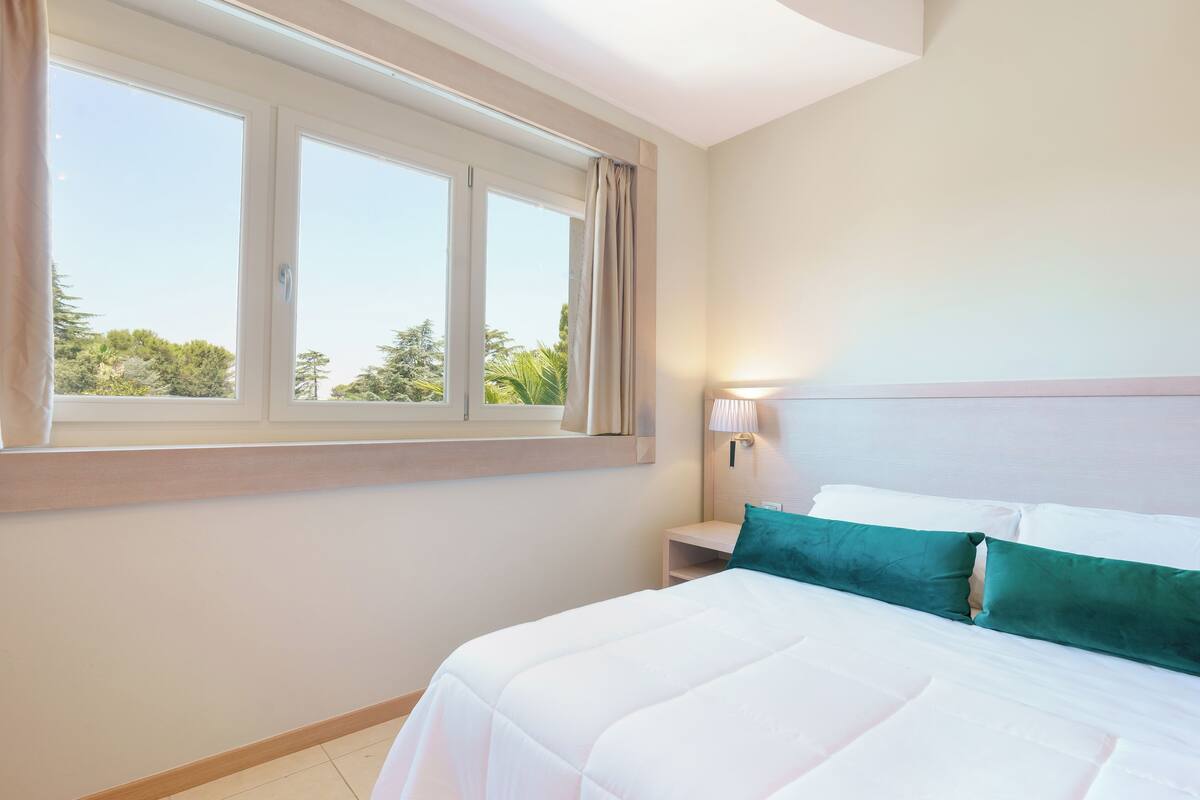 Et hotelværelse med et stort vindue, en seng med hvide sengetøj og en teal farvet pude, et natbord med en lampe, og udsigt til grøn vegetation udenfor.