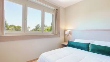 Een hotelkamer met een groot raam, een bed met witte beddengoed en een turquoise kussen, een nachtkastje met een lamp, en uitzicht op groen buiten.