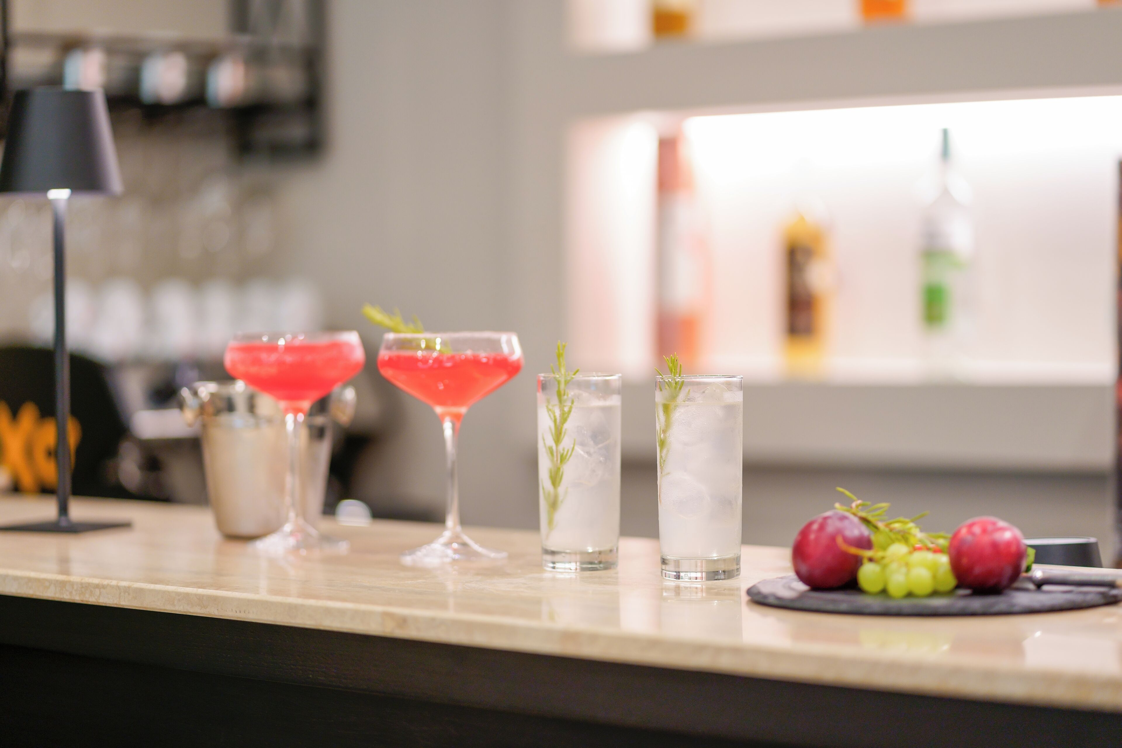 A bar area with a variety of drinks, including a red cocktail, a martini, and two tall glasses with herbs. There are also grapes and a small pitcher on the counter.