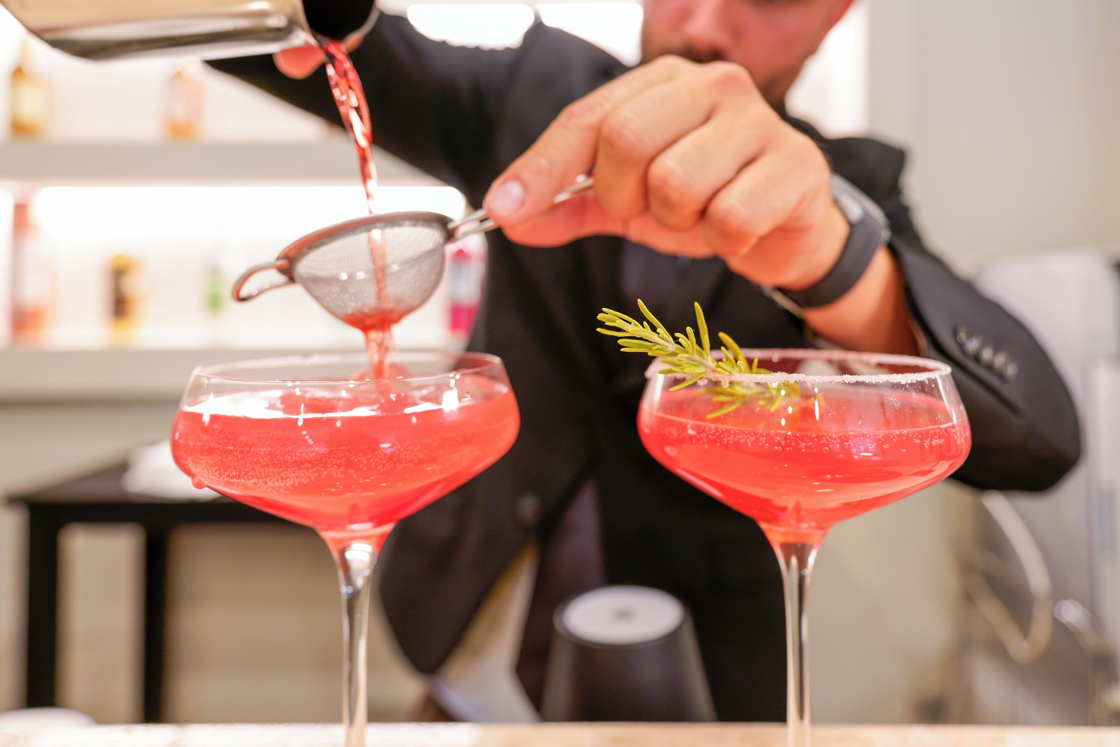 Un barman verse un cocktail rouge dans un verre à martini, en y ajoutant une branche de romarin.