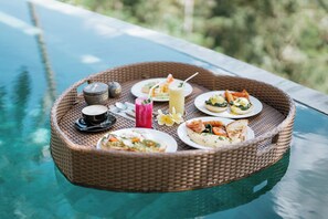 Ein Korbtablett mit verschiedenen Speisen und Getränken auf einem Glastisch am Pool.