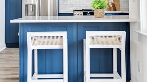 A kitchen with dark blue cabinets, a stainless steel refrigerator, a microwave, and a white island with two wicker barstools.