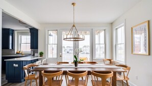 A spacious dining area with a large wooden table and six chairs, a chandelier, and a view of the city through large windows.