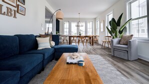 A modern living room with a blue sofa, a grey armchair, and a wooden coffee table. There is a dining area with a table and chairs in the background.