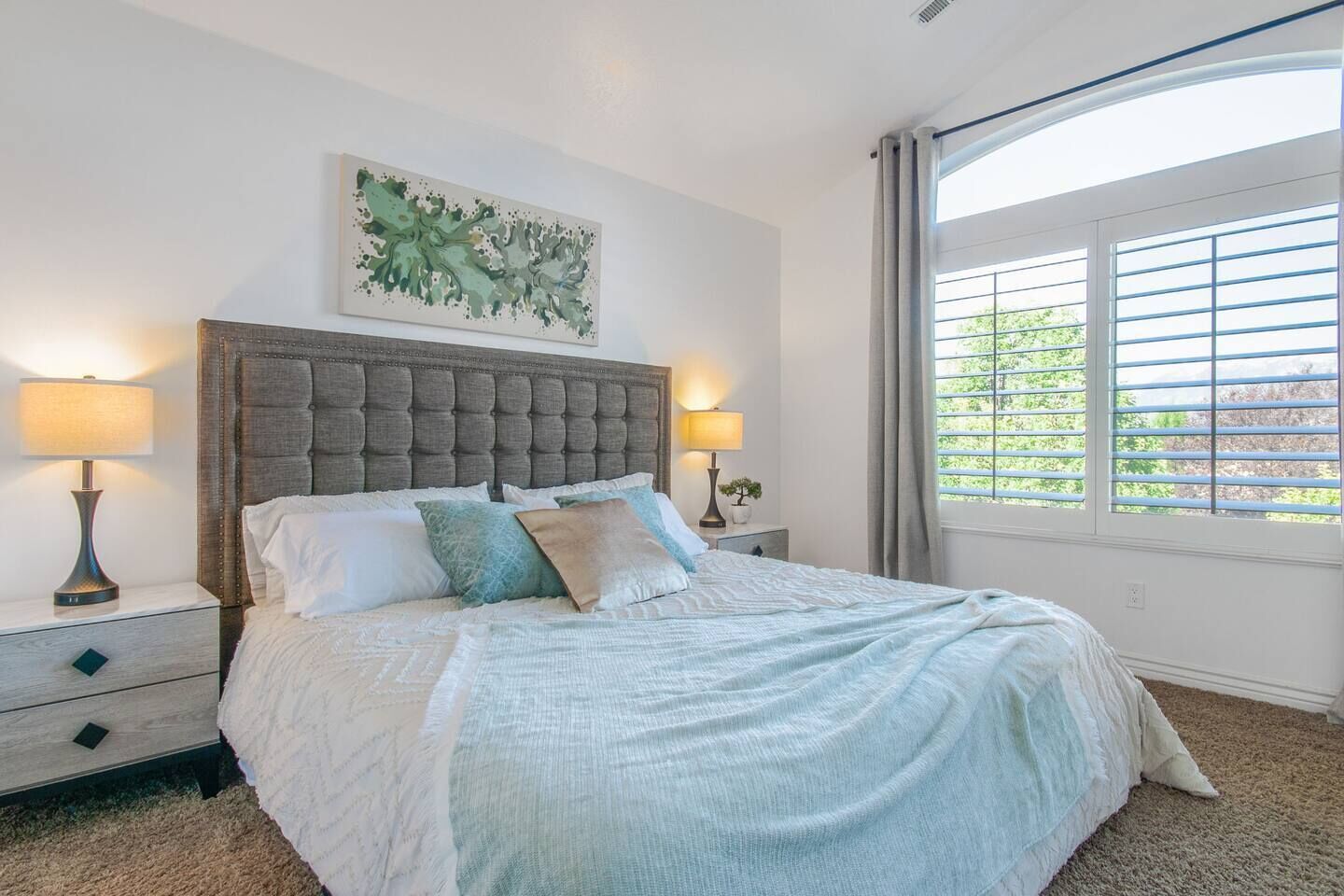 Quarto com uma cama grande, duas mesas de cabeceira, cabeceira, janela com persianas e uma obra de arte na parede.