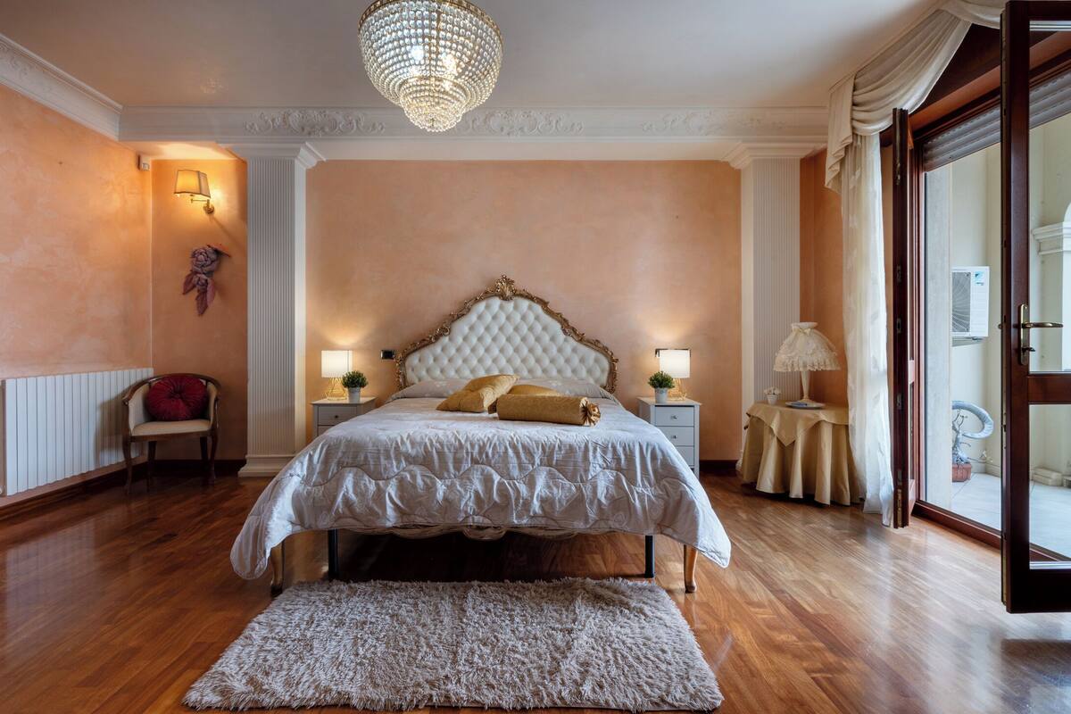 Une chambre avec un grand lit, deux tables de chevet avec des lampes, un lustre, un fauteuil rouge et une vue à travers une porte vitrée.