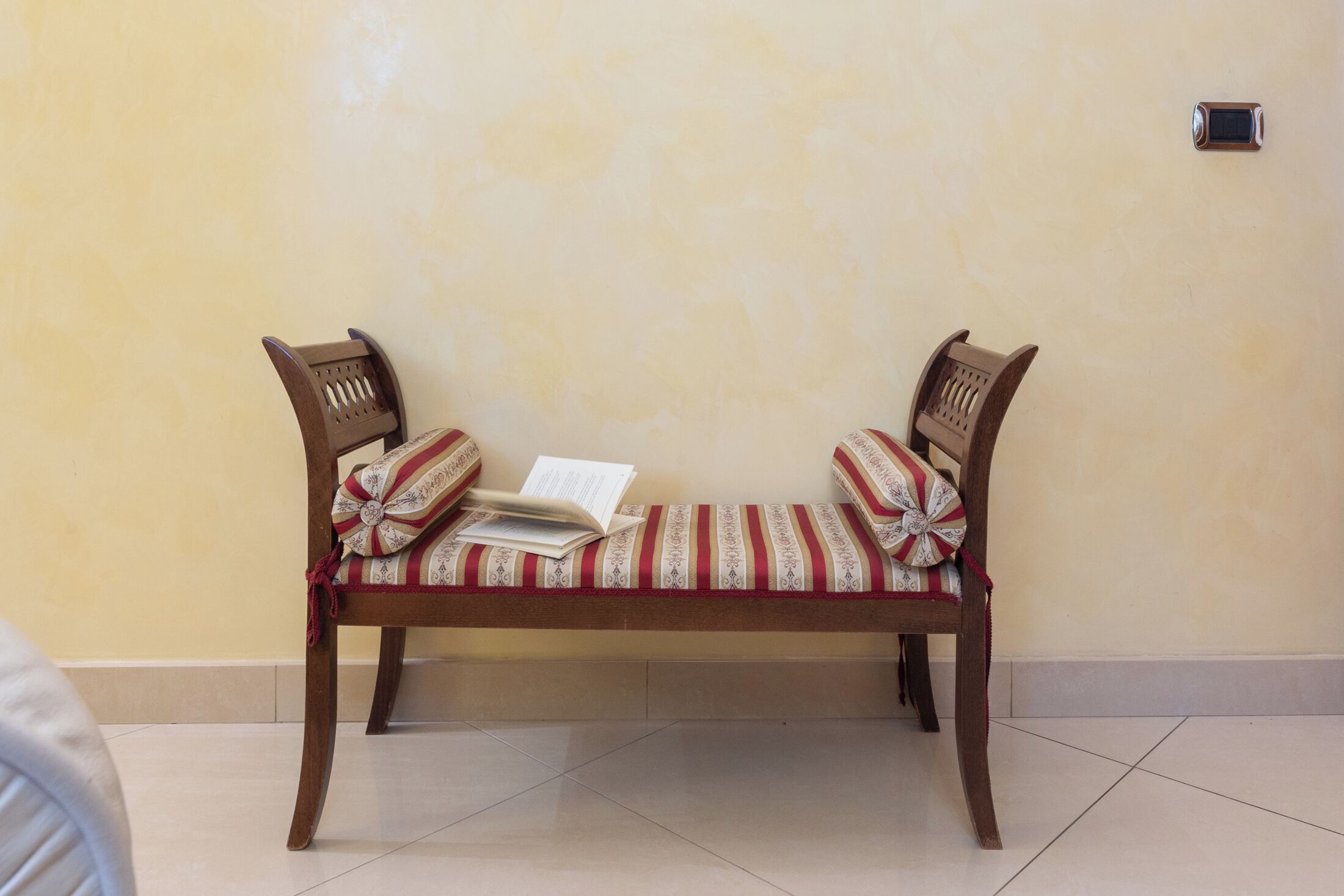 A wooden bench with striped cushions and a book on it, against a plain wall.