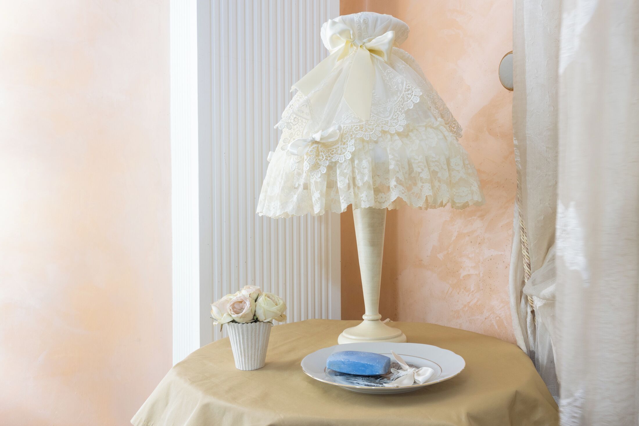 A room with a table set for tea, featuring a lace tablecloth, a white lamp, a plate with a blue soap bar, and a small vase with white flowers.
