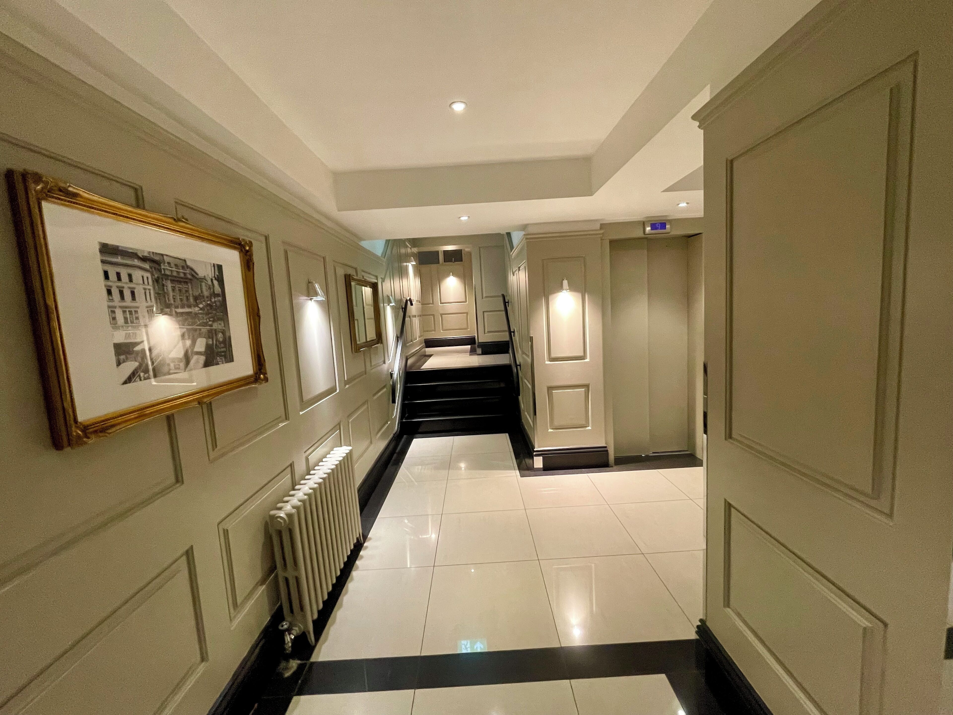 A hallway with a tiled floor, a radiator, framed pictures on the wall, and a staircase leading to an elevator.