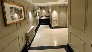 A hallway with a tiled floor, a radiator, framed pictures on the wall, and a staircase leading to an elevator. - Stylish studio/ Harrods /South Kensington /Chelsea/Central London (London)