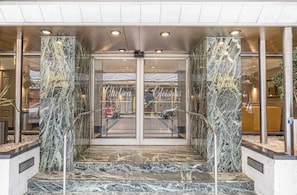 Entrada de un hotel con columnas de mármol, puertas de cristal y una escalera de metal.
