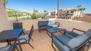 Ein Balkon mit einer Couch, Stühlen und einem Tisch, mit Blick auf ein Wohngebiet mit Gebäuden und Grünflächen.