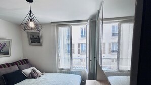 Un dormitorio con cama, ventana con vistas a edificios, radiador y lámpara colgante.